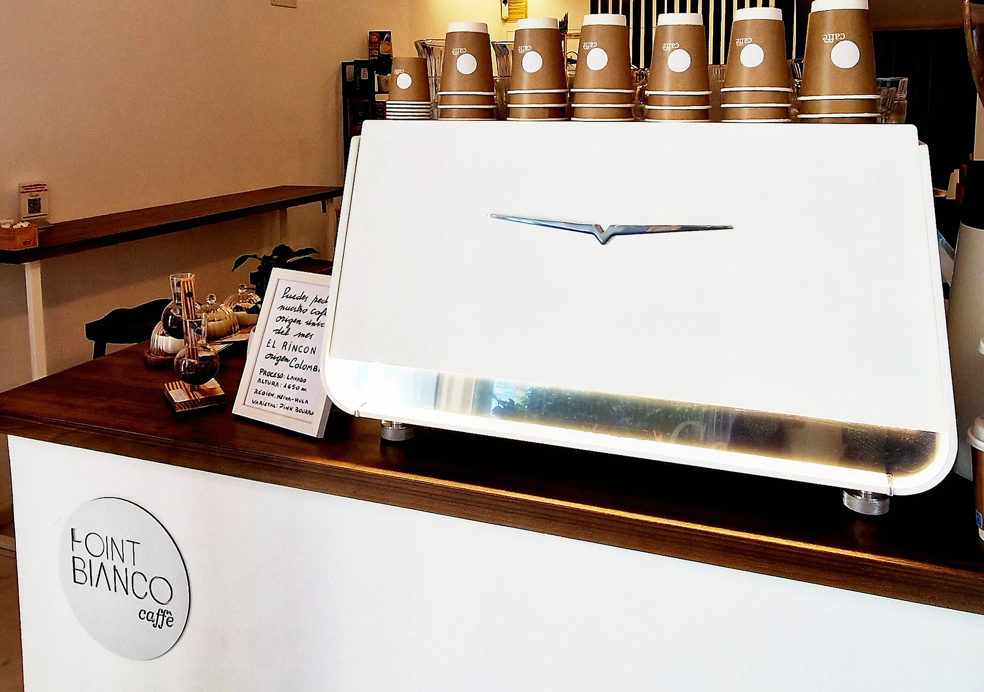 Barra blanca de Point Bianco con el logo en el frente, la cafetera Victoria Arduino, vasos de cartón apilados y lámparas de cristal