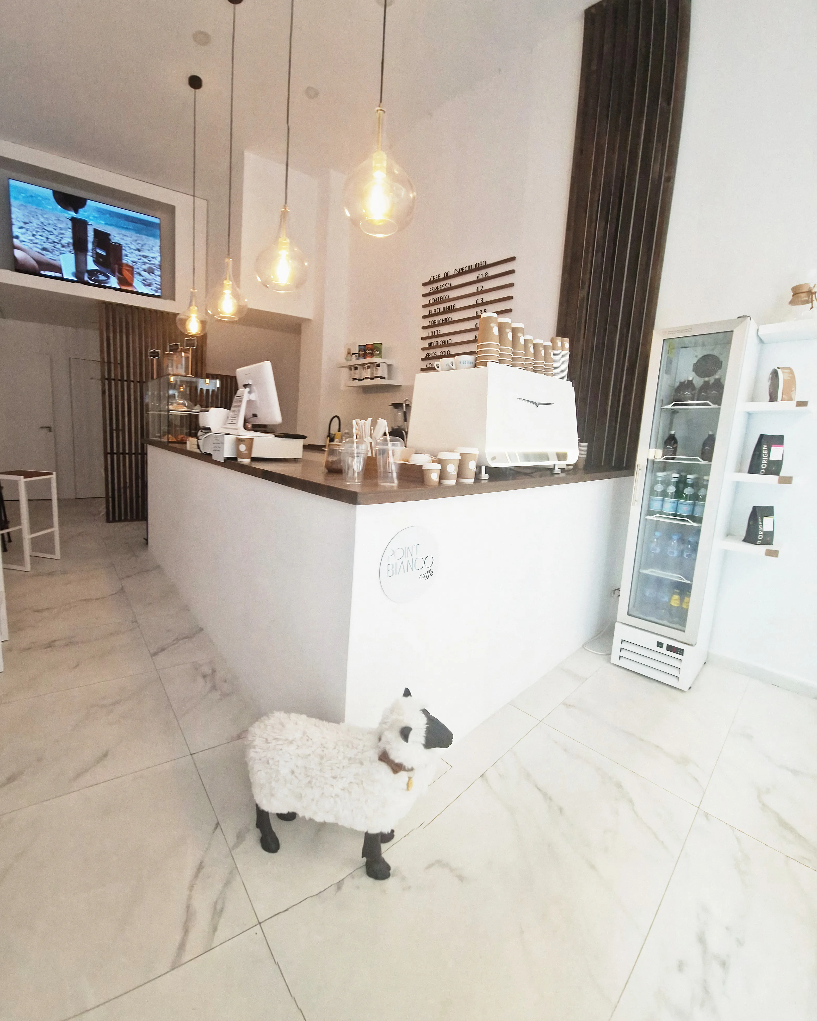 Interior de Point Bianco: suelo de mármol, la barra blanca con la cafetera, lámparas colgantes y una oveja decorativa junto a la barra